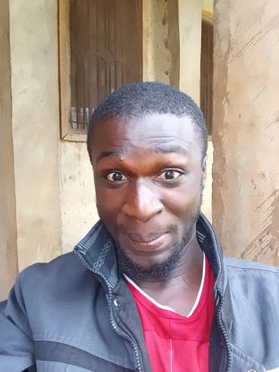 The image is a portrait of a young Black man, taken from the chest up, with the camera oriented vertically. He is wearing a dark gray jacket over a red t-shirt with a white trim around the collar. His head is slightly tilted, and he is looking directly at the camera with a neutral expression. He has short, dark, curly hair and a short beard. The background appears to be a textured, light brown wall with a wooden window frame to his right. The lighting suggests it is daytime, possibly indoors or in a shaded outdoor area. The image has a somewhat low resolution, and there is no visible text. The overall scene suggests a casual, everyday moment.