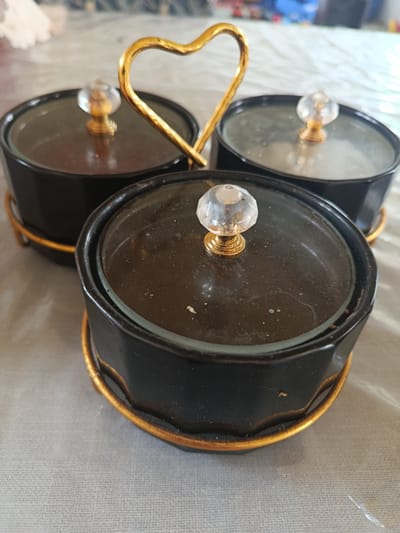 This media file shows a close-up, eye-level shot of a three-compartment serving dish. The dish is made of black material with gold accents, including a heart-shaped handle connecting the compartments. Each compartment has a clear glass lid topped with a crystal-like knob and a gold base. The dish sits on a gold wire rack. The background is blurred but appears to be a light-colored, textured surface, possibly a tablecloth, with hints of other objects and colors in the distance. There is no visible text. The lighting suggests it is daytime, likely indoors. The scene is static, with no activity taking place, focusing solely on the decorative serving dish. The setting appears to be a domestic or dining environment.