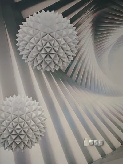 The image shows a close-up of a wall with a textured, abstract design. Two spherical, spiky objects, resembling geodesic domes or sea urchins, are prominently featured. The background has a swirling, pleated pattern in shades of gray and white, creating a sense of depth and movement. In the lower right portion of the image, a triple electrical outlet with a light switch is visible, mounted on the wall. The overall impression is of a modern, artistic interior design. The location specified, Гарочыцкі сельскі Савет, Belarus, suggests this is likely an interior space within a building in that rural administrative area. There are no people or discernible actions occurring in the image.