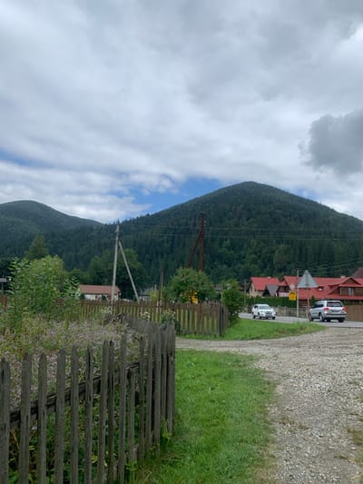 The image depicts a rural mountainous landscape in Tatariv, Ukraine, under an overcast sky with visible patches of blue. In the immediate foreground, a weathered wooden picket fence, partially broken, curves across the lower left, enclosing an area of overgrown vegetation with small purple flowering plants. To the right of the fence, a patch of green grass transitions into a light-colored gravel path or driveway. The midground features a paved road with two vehicles: a white crossover (Nissan Qashqai type) traveling away from the viewer on the left lane, and a silver SUV (likely a Toyota Highlander) further down the road, also moving away. Along the right side of the road, several residential structures with orange-red gabled roofs are visible. On the left midground, a light-colored utility pole with power lines stands, and a single-story house with a brown roof is partially visible behind vegetation. A darker utility pole is positioned further right. The background is dominated by two large, densely forested mountains, appearing to be covered predominantly by coniferous trees. No individuals are visible.