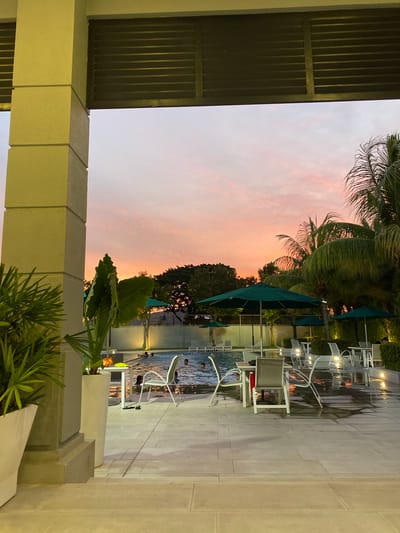 The image portrays an outdoor swimming pool and patio area at dusk in La Aurora, Ecuador. The sky displays distinct pink, orange, and light blue hues characteristic of a sunset, with a low cloud cover.
In the immediate foreground on the left, a large, light-colored architectural column stands vertically. To its left, a white planter contains green, palm-like foliage. Between the column and the pool, another tall white planter holds a large-leafed green plant.
The central feature is a rectangular swimming pool, actively occupied by approximately six to eight individuals who are partially submerged, appearing to be swimming or wading. The water surface reflects the ambient sky and artificial lighting. Surrounding the pool is a light-colored tiled patio, which exhibits wet patches from splashing or recent moisture.
Arranged on the patio are multiple dark green patio umbrellas, alongside numerous tables and chairs with light-colored frames. One table in the foreground has a yellow object and an orange object placed on its surface. A red garment is draped over a chair near another table. At the far end of the pool, several lounge chairs are visible.
The background features dense green vegetation, including numerous prominent palm trees on the right side, and a line of other trees against the horizon. A low wall or fence, partially illuminated by artificial lights, separates the pool area from the distant vegetation. Artificial lighting sources are active around the pool deck and the wall, providing illumination as natural light diminishes.