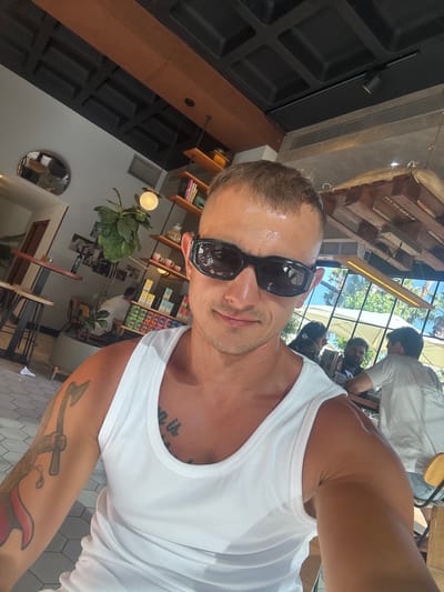 The image shows a man in a white tank top and sunglasses taking a selfie in what appears to be an indoor-outdoor cafe or restaurant. The man has short hair and a tattoo on his left arm. In the background, there are other patrons sitting at tables, some with umbrellas providing shade. The environment includes wooden accents, shelving with products, and large windows looking out onto a sunny, possibly tropical, scene. The cafe's ceiling has a unique design with dark panels and wooden beams. The flooring is tiled. The overall impression is a relaxed, casual setting.