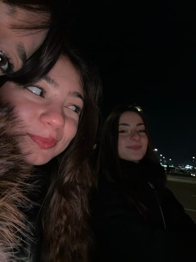 Two young women pose in a dark outdoor setting, illuminated by city lights faintly visible in the upper part of the frame. The woman in the foreground wears a fur-trimmed jacket and looks upwards with a slight smile. The other woman is in the upper right, also smiling, and wearing a dark jacket. The overall image has a casual, informal snapshot quality.