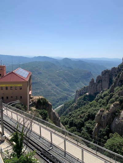 The image captures a view from the Montserrat mountain range in Catalonia, Spain, specifically near the municipality of Collbató. In the foreground, a building with a red-tiled roof, fitted with solar panels, is visible on the left. Below the building, a set of train tracks runs parallel to a pedestrian walkway with a metal railing. The background is dominated by a vast expanse of rolling green mountains and valleys, with a clear blue sky overhead. The terrain is rugged, with steep cliffs and dense vegetation. A winding road can be seen in the distance through one of the valleys. The scene is bathed in bright sunlight, suggesting a clear, sunny day.
