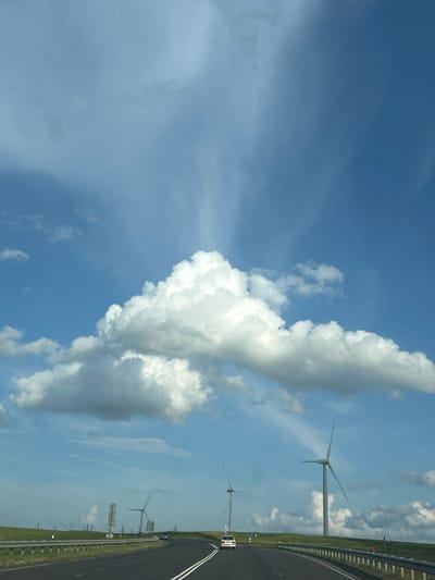 The image shows a view from a road, likely a highway, with a landscape featuring wind turbines. The road is paved and has double white lines indicating lanes. A silver car is visible on the road, driving away from the viewer. On the left side of the road, there is a metal guardrail and a grassy embankment leading to more wind turbines and some signage. The sky is blue with scattered white clouds, and there are rays of sunlight filtering through. The overall scene suggests an open, possibly rural area in the vicinity of Ладзеніцкі сельскі Савет, Belarus, characterized by renewable energy infrastructure.