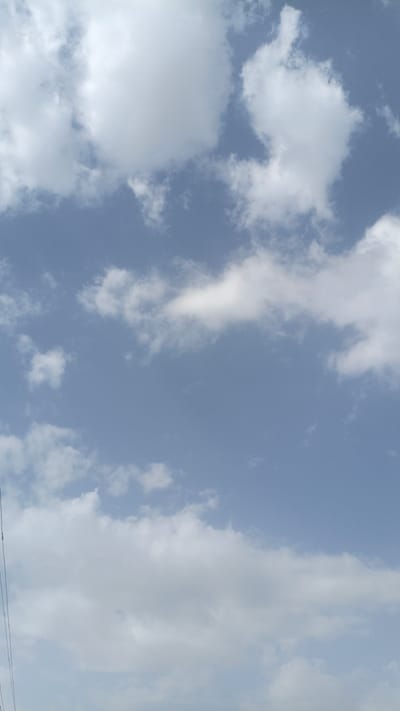 The image is a vertical, medium-angle shot of a cloudy sky, possibly in Нуъмон Розиқ, Tajikistan. The sky is a pale blue, with scattered white cumulus clouds of varying sizes and densities. The clouds appear to be in motion, with some appearing soft and wispy, while others are more defined and solid. A thin, dark, vertical line, potentially a wire or antenna, is visible on the far left of the frame, extending from the bottom edge upwards. No people or other discernible objects are present in the frame, suggesting an outdoor, possibly aerial, perspective.