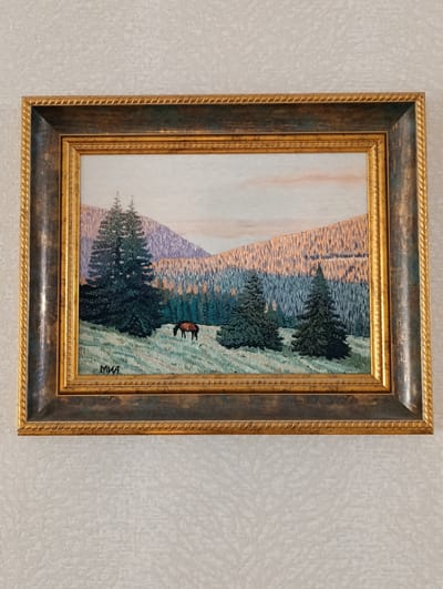 A framed landscape painting hangs on a light-colored, textured wall. The painting depicts a mountain scene with evergreen trees in the foreground and mountains in the background. A single brown horse is grazing in a grassy field. The sky shows hints of pink and orange. The painting is signed "MIA" in the lower left corner. The frame is ornate, with a gold rope-like pattern around the edges and a darker, possibly wood-grain finish on the broader part of the frame. There is no indication of the location as Barvinok, Ukraine in the image.