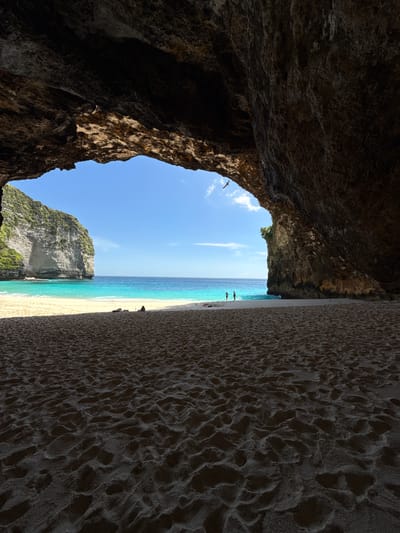 A vertical, medium shot captures a beach scene from within a cavernous opening, likely on the island of Nusa Penida, Indonesia. The perspective is set against a blue sky with a few wispy clouds. The right side of the frame features a sandy beach with patterned, ridged sand extending upwards. The turquoise and azure waters of the ocean form a vertical line between the beach on the right and a jagged rock formation with green foliage on the left. Two indistinct figures stand near the shoreline. The left side of the image is dominated by the dark, shadowed interior of the cave entrance. A single bird flies through the opening.
