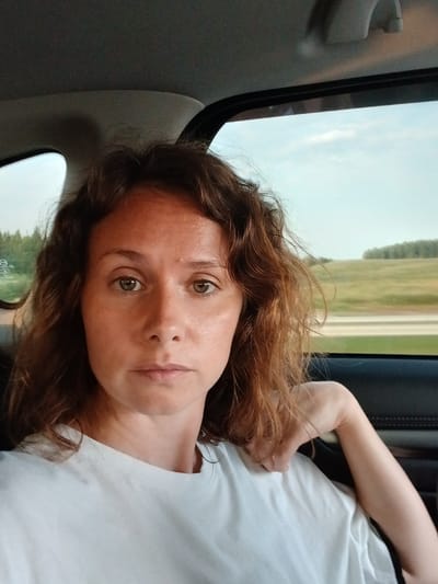 A woman with shoulder-length brown hair and freckles on her face is sitting in the back seat of a car. She is wearing a white t-shirt and her left arm is resting on the back of the seat. The background outside the car window shows a blurry landscape of green fields and trees under a cloudy sky. The image appears to be a selfie taken inside a moving vehicle. The location is identified as Novye Tatyshly, Russia.