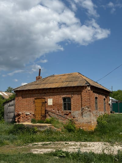 The image depicts a two-story brick building in Полянки, Russia. The building has a weathered, brown roof and a small chimney. The brick facade shows signs of disrepair with missing bricks. A wooden door is visible on the first floor, along with a sign. Windows are seen on both floors, some with ornamental grilles. The surrounding environment includes a gravel path, grass, and foliage, with a cloudy sky overhead.