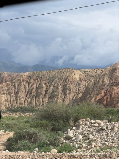 The image depicts a scenic view of the Fodor Valley near Tong, Kyrgyzstan. In the foreground, a rocky, arid terrain is visible, with a path leading into lush green vegetation. The middle ground is dominated by a large, eroded mountain range, displaying layers of sediment and sparse greenery. The background reveals a chain of distant, mist-covered mountains. The sky is overcast with dramatic clouds. There are no people or animals visible in the image. The overall scene conveys a sense of natural beauty and rugged landscape.
