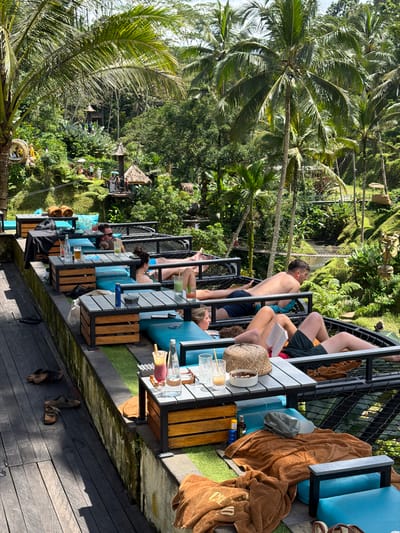 The image depicts a row of elevated seating platforms with people relaxing and enjoying drinks at a location in Tegal Suci, Indonesia. The platforms are equipped with blue cushioned chairs and tables set with glasses, bottles, and personal items. Palm trees and lush greenery surround the area, suggesting a tropical environment. Some individuals are sunbathing or reading, while others appear to be conversing. A small structure, possibly a bar or entertainment stage, is visible in the distance amidst the dense vegetation.