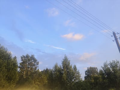 The image captures an outdoor scene with a clear blue sky dotted with a few wispy clouds. Several parallel power lines stretch diagonally across the upper right portion of the frame, connected to a utility pole on the far right. Below the sky, a dense line of green trees forms the horizon, with varying heights and leaf densities. The foliage appears lush and suggests a natural, possibly rural environment. In the foreground, a hazy, yellowish mist or light filters upwards from the bottom edge, obscuring the lower parts of the trees. The overall impression is a tranquil, natural setting under a clear sky, possibly during early morning or late afternoon given the quality of light. The location is identified as Kruglitski sielski Savek, Belarus. There are no people or animals visible in the image.