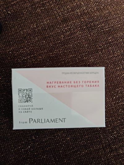 A pack of Parliament cigarettes is placed on a brown textured surface. The pack is white with pink and grey accents. Text on the pack reads "НАГРЕВАНИЕ БЕЗ ГОРЕНИЯ ВКУС НАСТОЯЩЕГО ТАБАКА" which translates to "HEATING WITHOUT BURNING THE TASTE OF REAL TOBACCO." There is also a QR code and the text "СКАНИРУЙ И УЗНАЙ БОЛЬШЕ НА САЙТЕ" which means "SCAN AND LEARN MORE ON THE WEBSITE." Below this is the word "from" and the Parliament logo. The image is a close-up and does not show the surrounding environment or any indication of its location in Klenovo, Russia.