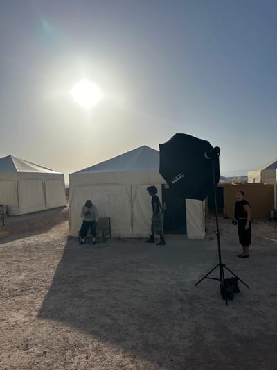The image depicts an outdoor setting with bright sunlight. There are several people present: one standing in the background dressed in all black, another standing near a white tent, and a third person sitting on a chair in front of a tent. The scene includes white tents, indicating a possible event or encampment. A large black Profoto softbox is prominently positioned on a stand. The location is Sidi Badhaj, Morocco, suggested by the terrain and architectural style.