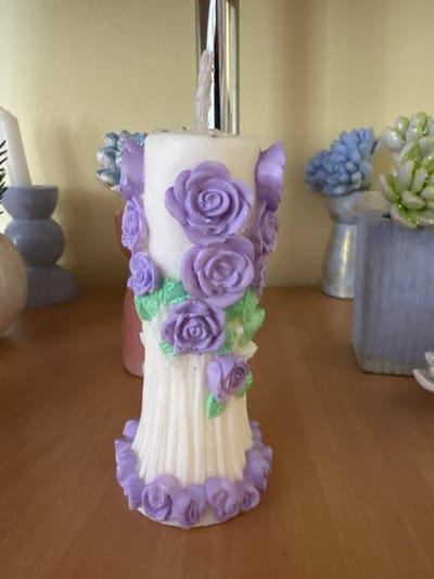 This is a close-up, eye-level, indoor shot of a decorative white candle adorned with purple rosebuds and green leaves. The candle has a ribbed texture and sits on a wooden surface. In the background, other decorative items are visible, including blue and green floral-like ornaments in vases, and a grey stackable candle holder with a white candle. The wall behind the objects is a light yellow. The scene is located in an indoor setting in Ветрынскі сельскі Савет, Belarus.