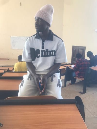 The image depicts an indoor classroom setting in Durumi, Nigeria. In the foreground, a male subject is positioned, wearing a light pink beanie with "LOEWE" embroidered, a grey polo shirt with a black collar and "Denim" printed across the chest, and light grey sweatpants featuring large dark letters, notably "ATTA" and "TA". He looks towards the right side of the frame, holding a dark smartphone vertically in his clasped hands.
Behind the subject, multiple rows of wooden desks with dark metal frames are arranged. On the left mid-ground, a person wearing a yellow shirt is partially visible, seated at a desk. On the right mid-ground, two individuals are seated at desks: one wears a red and black plaid shirt and appears to be engaged with papers on the desk, while the other wears dark clothing and headwear. The background wall is pale yellow, featuring a whiteboard with handwritten text on the left and a dark brown wooden door with two panels on the right. The floor is light-toned.