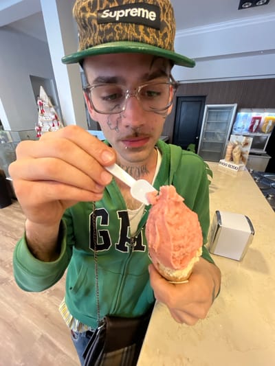 A person wearing a Supreme hat, glasses, and a green hoodie holds an ice cream cone with a small, white spoon. The ice cream is mostly pink and sits on top of a white cone. The person has facial tattoos. Behind them is a counter with a napkin dispenser, a freezer with what appears to be baked goods, and a wall. The picture is taken at an angle, making the scene appear tilted.