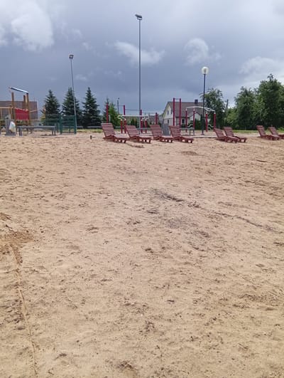 The image shows a sandy outdoor area, likely a beach or playground, in Šaukėnai, Lithuania. In the foreground, the sand is undisturbed, with small pebbles and indentations visible. In the mid-ground, several wooden lounge chairs are arranged on the sand. Behind the chairs, there is a playground structure with red and yellow elements, along with a metal fence and trees. In the background, there are residential buildings and more trees under a cloudy sky. Tall streetlights are also present. The scene depicts a public recreational space.