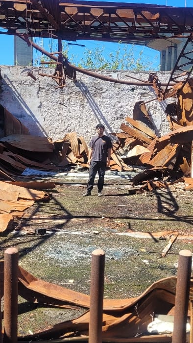 The image presents the interior of a significantly dilapidated or partially demolished industrial structure in Zárate, Argentina, under clear daylight conditions. A single male subject stands centrally in the mid-ground, facing the camera directly. He is dressed in a dark short-sleeved t-shirt with a small graphic on the left chest, dark trousers, and dark shoes. The ground is an uneven, dark, and possibly organic surface, strewn with rust-colored debris. Two vertical, cylindrical, heavily rusted metal posts are prominently positioned in the immediate foreground. The background consists of a tall, light-colored, textured wall exhibiting extensive damage and exposed surfaces. Large accumulations of heavily rusted, corrugated metal sheets are piled against this back wall, to both the left and right of the male subject, with additional sheets scattered across the floor. The original roof structure is largely absent, revealing a clear blue sky. Remnants of a rusted metal truss system span the upper portion of the frame. Beyond the damaged structure, green foliage and a large, light-colored concrete bridge or industrial facility are visible against the sky in the upper background. Strong sunlight illuminates the scene, casting distinct shadows from the subject and various debris elements onto the ground.