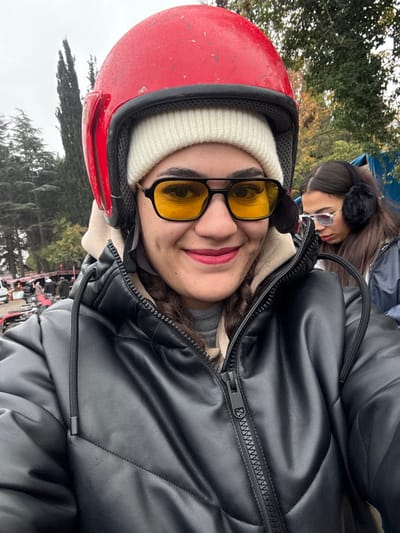 The image captures an outdoor scene in Çiftlikköy, Turkey, under an overcast sky. In the foreground, a female subject, appearing to take a selfie, is prominently featured. She wears a red full-face helmet over a white knit beanie, yellow-tinted sunglasses with a black frame, and a black faux leather puffer jacket with a visible zipper, worn over a beige layer. Her lips are covered in dark pink lipstick, and she displays a smile with visible dimples. To the right and slightly behind her, a second female is partially visible, wearing black fluffy earmuffs and light-framed, lightly tinted sunglasses, looking downwards and to the right. The background features tall, dark green coniferous trees, dense green foliage, and several all-terrain vehicles (ATVs) with individuals gathered around them, indicative of an ATV riding or rental area. Red and white striped barrier tape is discernible in the left background near the ATVs. A blue tarpaulin or structure is faintly visible on the far right side.