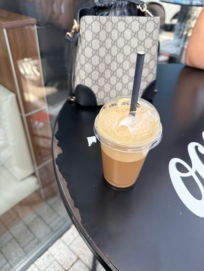 In Chuchuligovo, Bulgaria, on a black, circular table with a distressed edge, sits a clear plastic cup filled with a frothy, light brown beverage, topped with a domed lid. A dark gray paper straw extends from the center of the lid. Behind the cup, a beige handbag with a repeating "GG" logo pattern and black trim is visible. The background shows a reflective surface that appears to be a glass window, with a wooden structure to the left and hints of a car and building beyond. White lettering, partially obscured, is present on the table to the right.