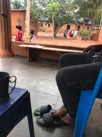 The image depicts a person sitting in a blue plastic chair on a tiled floor. The person is wearing dark gray jeans and gray quilted slides. A black mug sits on a blue table next to the chair. Through a window, a scene outside shows children playing in a dusty yard surrounded by orange-colored buildings and trees. In the background, a group of people is walking down a dirt path. The scene is located in Otukpa, Nigeria.