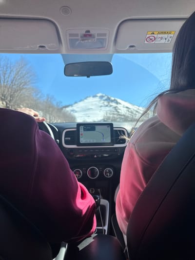 The image depicts an interior view from the rear passenger compartment of a vehicle, looking forward. Two individuals occupy the front seats. The driver, on the left, wears a dark red long-sleeved top, with their left hand visible on the steering wheel. The front passenger, on the right, is seen from behind, with dark hair and wearing a light pink long-sleeved top; a black seatbelt strap crosses their right shoulder.

The vehicle's dark dashboard features a centrally mounted infotainment screen displaying a navigation map with a blue route. Below the screen are circular controls and a gear shifter with a visible USB port. A rear-view mirror is affixed to the windshield, and an overhead console contains interior lighting and a red-triangle button, possibly for emergency services or hazards. The passenger-side sun visor is visible, bearing an "AIRBAG" warning label.

Through the clear front windshield, a prominent, snow-covered mountain peak is visible in the distance, dominating the landscape under a clear blue sky. Sparse, bare trees line the apparent roadside on either side of the vehicle. Bright sunlight illuminates the scene, casting distinct shadows within the vehicle's interior. The scene is set in Gradačka Poljana, Montenegro, characterized by a mountainous, winter environment.