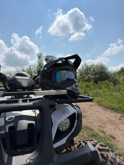 This is a close-up, low-angle shot of a black helmet with rainbow-tinted goggles resting on the front rack of a white ATV. The ATV is parked on a dirt path that curves to the right, bordered by green grass and dense foliage. In the background, the sky is a bright blue with fluffy white clouds and a faint contrail from an aircraft. Another helmet, this one white with a green "Monster Energy" logo, is visible behind the black helmet on the ATV's rack. The ATV's headlight is also visible, along with part of its tire. The scene appears to be set in a rural or outdoor recreational area, possibly a trail or park. The image is taken in Gushchino, Russia.