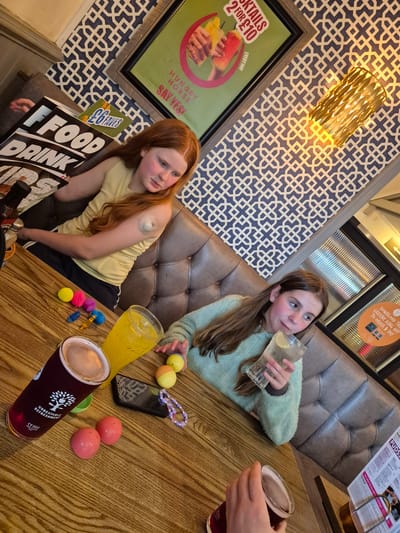 The image shows two young girls sitting at a table in what appears to be a pub or restaurant. The girl on the left has red hair and is wearing a yellow top and black shorts. She has a medical device attached to her arm. The girl on the right has brown hair and is wearing a light green sweater, and is drinking from a glass with lemon slices.
On the table, there are several objects including a smartphone, toy balls, a purple bead bracelet, and drinks in pint glasses. One drink is golden in color, the other is dark.
The background shows a padded booth seating area and patterned wallpaper with a framed advertisement for cocktails. There are also menus on the table. The OCR text includes "HUNGRY HORSE" which is a chain of pubs in the United Kingdom. This image was probably taken at a Hungry Horse pub. The OCR also has "GREENE KING APP" which is another chain of pubs in the United Kingdom. The image might be taken at a pub from either the Hungry Horse or Greene King chains.