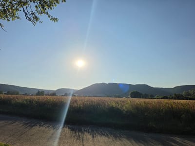The image shows a scenic view of a rural landscape under a bright, sunny sky. In the foreground, a paved road runs diagonally across the bottom of the frame, casting long shadows from an unseen object. To the right of the road is a field of tall, golden-brown grass, likely a crop such as wheat or corn, which extends towards rolling hills in the background. The sun is prominent in the upper center of the image, emitting a strong lens flare that creates a diagonal beam of light across the sky and into the field. The hills in the distance are covered in green foliage, with some scattered trees and structures suggesting a small village or settlement nestled amongst them. The sky is a clear, pale blue. The overall impression is one of a peaceful, sun-drenched rural setting. This scene is located in the vicinity of Godelheim, Germany.