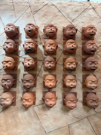 The image displays a textured, light brown stone wall with several rows of terracotta monkey head sculptures mounted on square terracotta tiles. The sculptures vary in expression, with some appearing to smile, laugh, or stick out their tongues. The lighting suggests an outdoor setting with natural light casting shadows on the wall and sculptures. The scene is located in Tambon Mae Raem, Thailand.