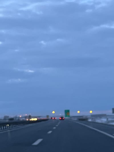 The image displays a blurry, low-light view from the interior of a vehicle traveling on a multi-lane highway, consistent with the A1 highway near Kovilj, Serbia. The upper two-thirds of the frame are dominated by an overcast, blue-gray sky exhibiting indistinct cloud formations. The lower third features the asphalt roadway, marked with white dashed lane lines.
Essential details include:
* **Environment:** The setting is an outdoor highway during what appears to be dusk, dawn, or under heavy overcast conditions. On the left side of the road, a metal guardrail runs parallel to the direction of travel. On the right side, beyond the road shoulder, indistinct white patches suggest the presence of snow or ice.
* **Objects/Subjects:**
* **Vehicles:** Multiple blurred red lights, indicative of vehicle taillights, are visible ahead in the distance, suggesting other traffic on the highway. Some blurred yellow/white lights are visible on the left side, possibly from oncoming traffic or roadside fixtures.
* **Road Signage:** A green highway sign, partially visible and blurred, stands on the right side of the road, likely indicating an exit or junction.
* **Lighting:** Three distinct, circular, yellow light sources, appearing as out-of-focus streetlights or indicator lights, are positioned above the roadway in the mid-ground.
* **Actions/Interactions:** The image implies forward motion from the perspective of the observer within a moving vehicle on the highway. No explicit human figures or specific interactions are discernible.