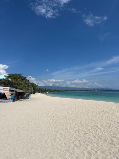 The image depicts a pristine white sand beach on a sunny day, likely in Saavedra, Philippines, based on the provided context. The foreground is dominated by the expansive sandy beach, showing subtle undulations and scattered small debris. In the middle ground, to the left, a small cluster of structures appears to be commercial establishments or stalls. One structure, identified by signage as "TANGUY LUNESA'S STORE," has a rustic appearance with wooden planks and a covered area. Blue chairs are visible outside this store. Beyond these structures, a line of lush green trees runs along the edge of the beach. A street lamp stands near the trees.
The turquoise water of the sea stretches out to the horizon, with a clear demarcation between the shallow, lighter blue near the shore and the deeper blue further out. Several small boats are scattered on the water. In the distance, a range of mountains forms the coastline, partially obscured by clouds. The sky is a vibrant blue, dotted with fluffy white clouds, some of which are thin and wispy, while others are more substantial cumulus formations. The overall atmosphere is tranquil and idyllic, characteristic of a tropical beach destination. A few indistinct figures are visible walking on the beach and swimming in the water.