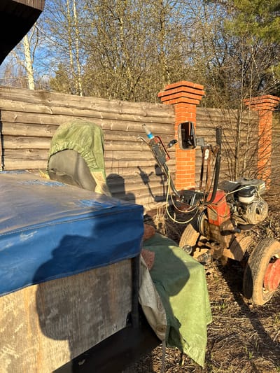 The image shows an outdoor scene, possibly a backyard or storage area, in Galevo, Russia. The environment appears rustic, with a wooden fence and ground covered in dirt and some vegetation.
Several objects are present: a gasoline-powered engine, possibly from a tiller or other small machine, rests on a small wheeled chassis, bricks, and what looks like a blue tarp or protective cover. There is also a smaller motorized machine.
No people or interactions are visible in the image.