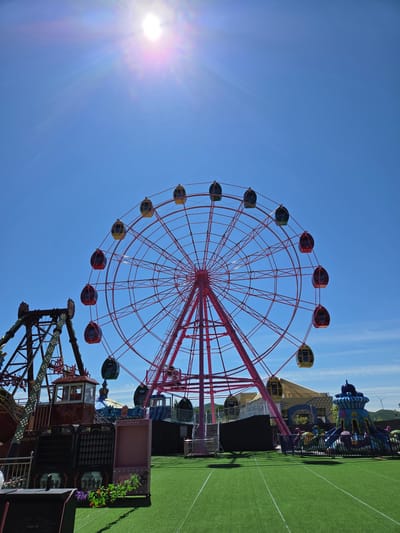 This image, taken in Jiejinkou, China, presents a vibrant amusement park scene under a clear, bright blue sky. The photo is oriented vertically, rotated 90 degrees clockwise.

The central and most prominent subject is a large Ferris wheel with a red or pink metal structure. Its many cabins are painted in alternating red, green, and yellow colors, adding splashes of contrast to the bright framework. The Ferris wheel dominates the center-left portion of the frame, reaching from the bottom towards the top and right.

To the far left, the bright sun is visible, casting a strong lens flare across the deep blue sky, indicating full daylight conditions. The sky occupies a significant portion of the image, particularly on the left side, and appears cloudless.

On the right side of the image, the ground is covered with artificial green turf, segmented by white lines, suggesting a recreational area within the park. Various other amusement park attractions and structures are partially visible in this area, including colorful, whimsical buildings or rides. Faintly in the far background, under the clear sky, distant hills or mountains can be seen, along with a small wind turbine, hinting at the surrounding landscape.

No specific activity involving people is depicted, but the scene clearly portrays an active amusement park setting designed for entertainment and leisure. The bright sunlight and clear sky suggest good weather.

Visible text on a dark structure near the base of the Ferris wheel on the lower right includes the Chinese characters "请勿" (qǐng wù), which typically translates to "Please do not..." likely part of a warning sign. Another panel shows a grid of small squares, possibly part of a game or display, but further text is illegible.