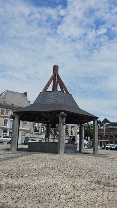 A well structure is visible in the center of the image, in the Place des Trois Fers in Philippeville, Belgium. The well structure has a dark grey, hexagonal roof with a wooden apex structure. The roof is supported by four thick, grey stone pillars. Inside the well structure, a grey stone basin is visible. There are two people sitting on the edge of the basin, one with blonde hair and the other with dark hair. They are facing away from the camera, towards the well. Behind the well structure, there are buildings with traditional architecture. The ground in front of the well is paved with cobblestones. The sky above is blue with scattered white clouds. The overall impression is of a peaceful town square on a cloudy day.