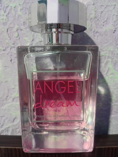 A square glass perfume bottle with a silver cap sits on a dark wooden surface. The bottle is filled with a pink liquid and has a label that reads "ANGEL dream" in pink font. Below this, it says "e100ml EAU DE PARFUM 3.4Fl.oz". The background is a textured purple and white wall. There is no discernible indication of the location in Bocani, Moldova.