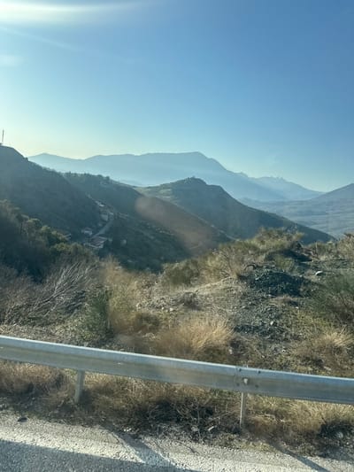 The image depicts a panoramic view of mountainous terrain in Μεγάλη Κερασιά, Greece, under a clear, bright blue sky with a prominent sun flare in the upper left. In the immediate foreground, a silver-colored metal guardrail spans horizontally across the lower frame, positioned above a light-colored paved road surface. Behind the guardrail, the ground slopes steeply downwards, characterized by dry, sparse, yellowish-brown vegetation, interspersed with rocky and gravelly patches.
The midground features multiple layers of green and brown hillsides descending into a valley. A narrow, winding paved road is visible traversing a lower hillside, with at least two small, light-colored buildings or houses situated along its path. On the leftmost peak of a foreground hill, a tall, slender lattice structure, consistent with a communication tower, is present.
In the background, successive layers of mountains appear in varying shades of blue due to atmospheric haze, extending towards the horizon. A distinct, taller peak with visible white or light-colored patches (potentially snow or bare rock) is discernible on the upper right side of the distant mountains. No individuals are visible in the scene; it is a static landscape captured in daylight.