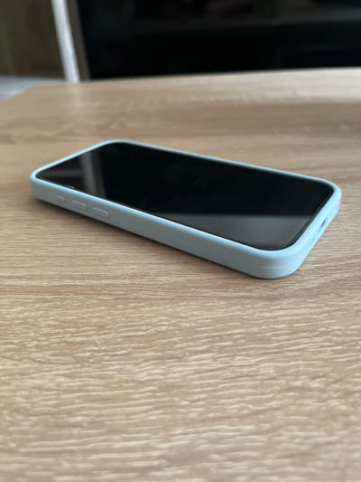 A light blue smartphone case encloses a smartphone with a black screen. The phone is lying on its side on a light brown wooden table, with the screen facing upwards. The table has a visible wood grain pattern. In the background, a dark, out-of-focus object, possibly a television or computer monitor, is visible. The scene appears to be indoors.