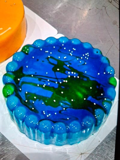 In Medvezhyi Ozyora, Russia, on a metallic surface, a round cake decorated to resemble the Earth sits on a white paper liner. The cake's icing is predominantly blue, with swirls of green representing continents. Tiny white dots are scattered across the blue to mimic stars. To the left of this cake, a portion of another round cake with orange icing is visible. The background shows a light-colored, slightly textured surface, possibly a table or counter, with some minor imperfections and what appear to be fine scratches or cracks.