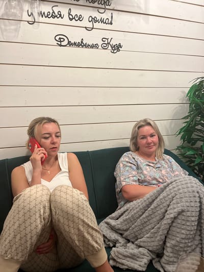 An interior scene in Kamyennoye, Russia, features two adult women seated on a dark green upholstered sofa against a white, horizontally paneled wall.
On the left, a woman with blonde hair, estimated to be in her 30s or 40s, is engaged in a phone call, holding a red smartphone to her right ear. She wears a white sleeveless top and light-colored sweatpants adorned with a repeating pattern resembling interlocking 'G's or similar monograms. A gold necklace with a small cross pendant is visible around her neck. Her expression is focused or serious.
To her right, a second woman, with blonde, shoulder-length hair and appearing to be in her late 40s or 50s, is seated, looking towards the viewer with a slight smile. She wears a light-colored short-sleeved top patterned with various pastel-colored cats (pink, blue, grey). Her lower body is covered by a grey, textured, plush blanket, under which her arms are crossed.
The white paneled wall behind them features black script-style wall decals. The upper visible text reads "у тебя все дома!" (u tebya vse doma!, translating to "Is everything okay at home?" or "Are you all there?"). Below this, the text "Домовенок Кузя" (Domovenok Kuzya) is visible, referring to a popular Russian cartoon character. A green leafy plant is situated in the background on the far right of the frame. The lighting is even, suggesting an indoor setting.