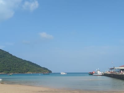A wide-angle landscape photograph depicts a coastal scene in the city of Preah Sihanouk, Cambodia. The view encompasses a sandy beach, calm ocean waters, a forested mountain, and a pier with docked vessels under a clear blue sky with sparse white clouds.

On the left side of the frame, a large, densely vegetated mountain slopes down to the water's edge. In the mid-ground, the turquoise ocean water is calm, reflecting the sky. Two small, traditional-style boats are visible near the base of the mountain. Further right in the mid-ground, a white yacht or speed boat traverses the open water.

The foreground features a light brown sandy beach that meets the shallow, clear water. To the right, a substantial pier extends into the water. Docked alongside the pier are at least two vessels: a prominent white and red passenger ferry or speed boat, and a darker-hulled boat partially obscured behind it. Structures with red roofs and open-air areas are built along the pier. Several individuals are visible on the pier, appearing to be walking or standing near the docked boats or structures. The overall environment suggests a bustling coastal area or ferry terminal.