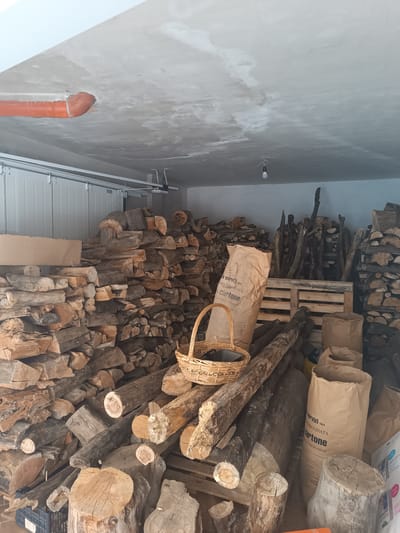 A dimly lit garage or storage area is filled with stacks of cut firewood. The firewood is of various sizes and shapes, with some logs appearing freshly cut and others more weathered. Several large paper bags, one with visible text that reads "a servizi DIFFERENZIATA Cartone," are placed amongst the wood. A woven basket containing some dark objects sits on top of a stack of logs. In the background, a section of a white garage door is visible, along with a single light bulb hanging from the ceiling. A large orange pipe protrudes from the concrete ceiling. The scene suggests a storage area for firewood, possibly for personal use or sale, and the time of day is likely daytime given the light entering the space. The overall impression is one of a functional, somewhat cluttered storage environment.