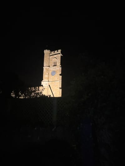 A low-angle shot depicts a lit medieval stone building at night. The structure features a clock and arched windows. Dark foliage surrounds the building, with a trellis-like fence visible on the right side. The building is lit with a warm glow and stands in stark contrast to the deep black sky and surrounding darkness.