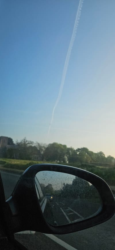 A view from a car window, featuring a side mirror and a bright sky. The side mirror is positioned in the right portion of the frame, reflecting a road and a blurred building or structure. The mirror's surface is speckled, possibly with water droplets or dirt. Outside the mirror, the scenery includes a stretch of green foliage and trees. Dominating the left portion of the image is a clear blue sky with a white contrail stretching across it. The scene suggests a journey or travel setting, possibly on a road trip in Broadland, United Kingdom. The time of day appears to be during daylight, with soft light illuminating the scene.