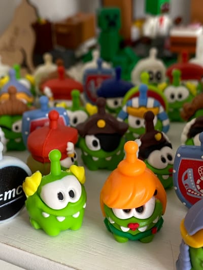 This image depicts a close-up, eye-level, medium shot of a collection of green, frog-like plastic toys, specifically Om Nom characters from the game Cut the Rope, arranged on a white shelf. The toys are in various colors and accessories, with some featuring orange hair, brown hats with yellow accents, or blue and white striped headwear. One toy on the left has yellow ear-like protrusions. In the background, out of focus, are other toys and objects, including what appears to be a green Minecraft Creeper figure, brown boxes, and possibly a light brown cardboard cutout of a cat. The lighting is soft and diffused, casting minimal shadows. There are no people visible in the image, and no discernible elements place the scene specifically in Ostrovki, Russia.