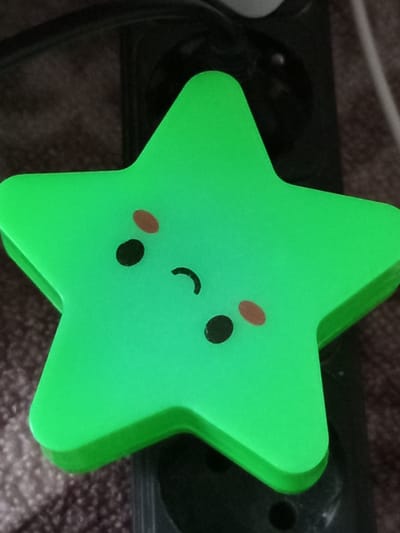 A bright green star-shaped object with a simple cartoon face is positioned on a dark, textured surface. The face consists of two black circular eyes, two small oval pinkish cheeks, and a curved black mouth. The object appears to be a decorative item or a light. The background is out of focus but suggests an indoor environment with dark surfaces and some lighter fibers. The lighting is somewhat dim, causing the green object to appear luminous. The location within the city of Älimbet, Kazakhstan, is not determinable from the visual information provided in the image.