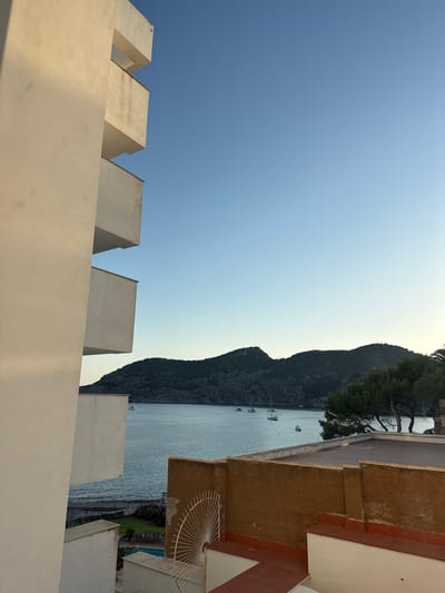 This is a vertically oriented, eye-level photograph taken from a balcony or rooftop overlooking a bay. The foreground features the textured, cream-colored stucco wall of a building with crenellated parapets. Beyond this, the calm, blue-gray water of a bay is visible, dotted with several sailboats. In the distance, a dark, tree-covered mountain slopes down to the water's edge. The sky above is a clear, pale blue, transitioning to a lighter hue near the horizon.

To the right of the frame, part of another building is visible, with walls of reddish-brown and white stucco. A decorative, white metal fan-shaped object is mounted on the reddish-brown wall. The overall impression is one of a serene coastal setting, likely in the late afternoon or early evening given the soft light and the calm water. The location cues, such as the architecture and the bay with sailboats, are consistent with a Mediterranean coastal town. There is no visible text in the image.