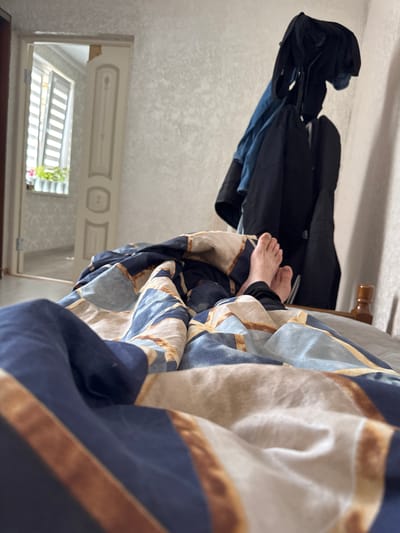 The image shows a person's bare feet emerging from under a duvet with a geometric pattern of blue, beige, and white. The person is lying in a bed in a room. In the background, a doorway is open, revealing a hallway and a window with blinds and potted plants. To the right of the doorway, a coat rack is covered with several dark jackets. The wall behind the coat rack is textured. The lighting suggests it is daytime. The image appears to be taken inside a residential dwelling in Nazran, Russia, likely a bedroom.